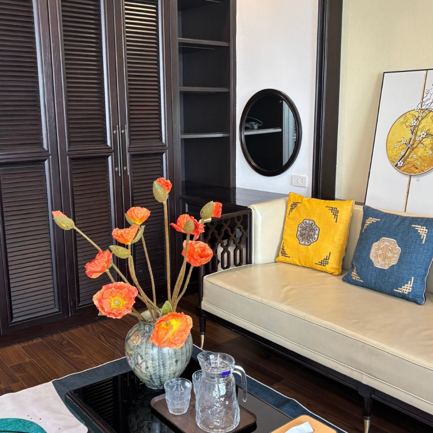 A modern living room with a sofa, decorative pillows, a vase with flowers, and a round mirror on the wall.