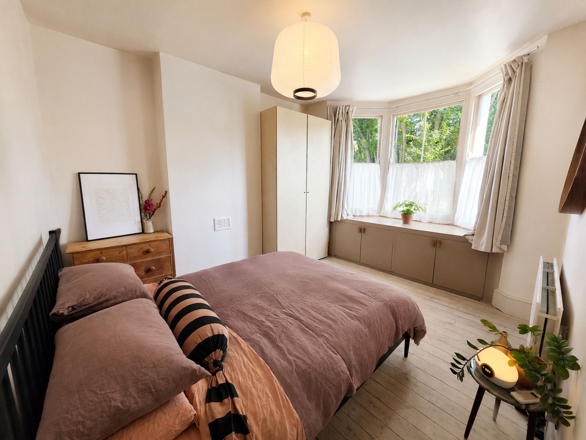 Un dormitorio con cama, mesita de noche, lámpara, ventana con cortinas y una planta en maceta.
