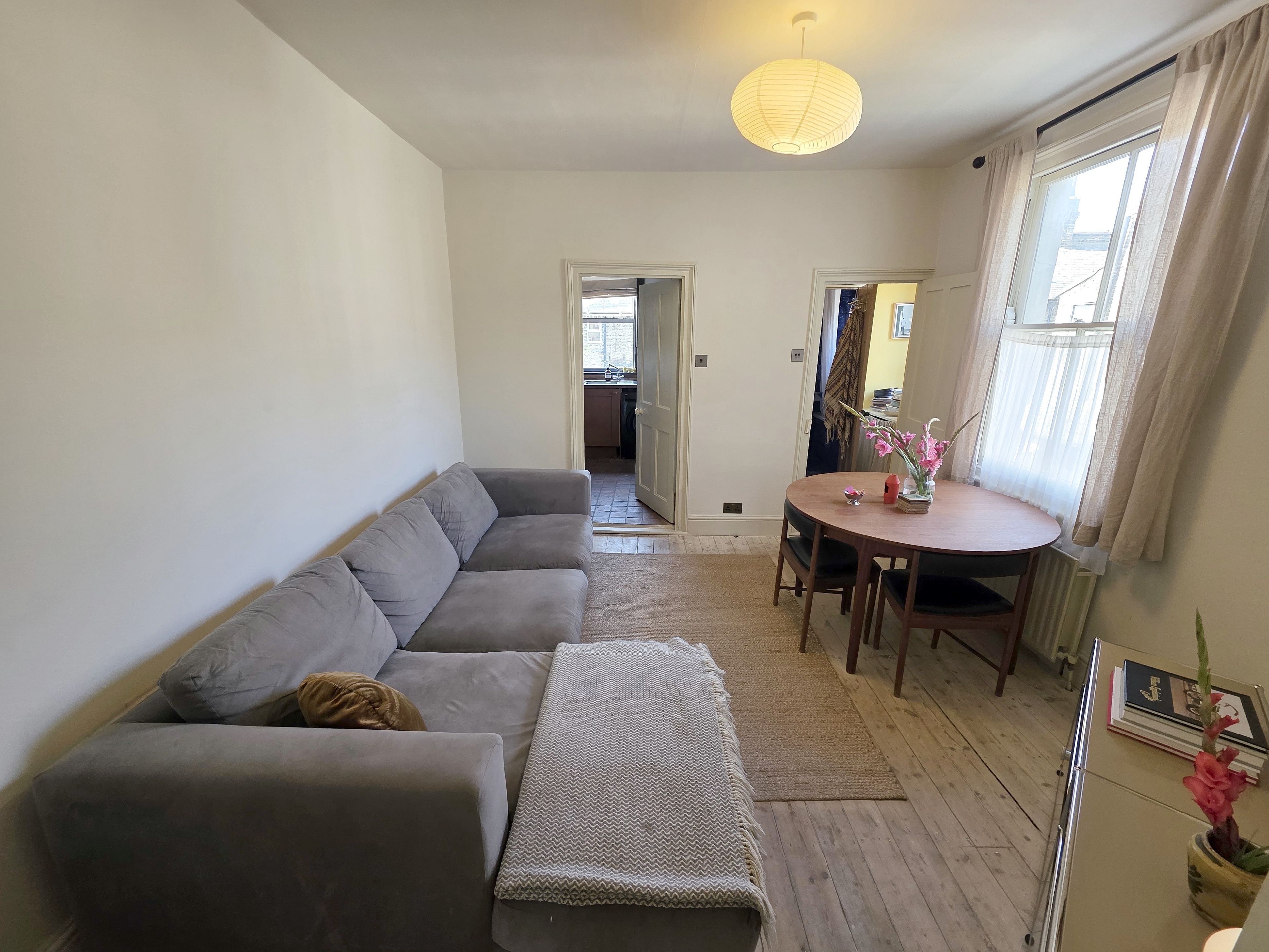 A living room with a grey sofa, a small round table with a vase of flowers, and a view through an open door to a kitchen area.