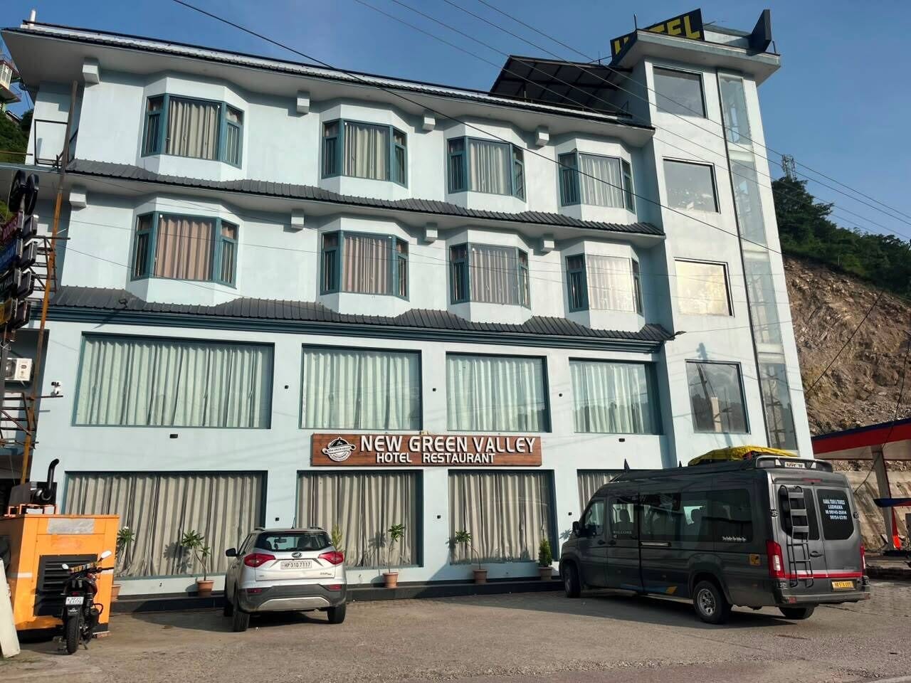 Un immeuble à plusieurs étages, sur lequel on peut lire l’inscription « New Green Valley Hotel Restaurant ». Deux voitures sont garées devant, et une camionnette est stationnée sur le côté droit.