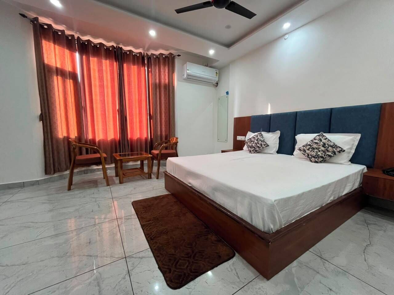 A hotel room with a bed, two chairs, a desk, a ceiling fan, and an air conditioning unit.