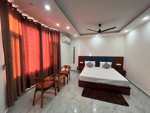 A hotel room with a bed, a desk, chairs, a ceiling fan, and an air conditioning unit.