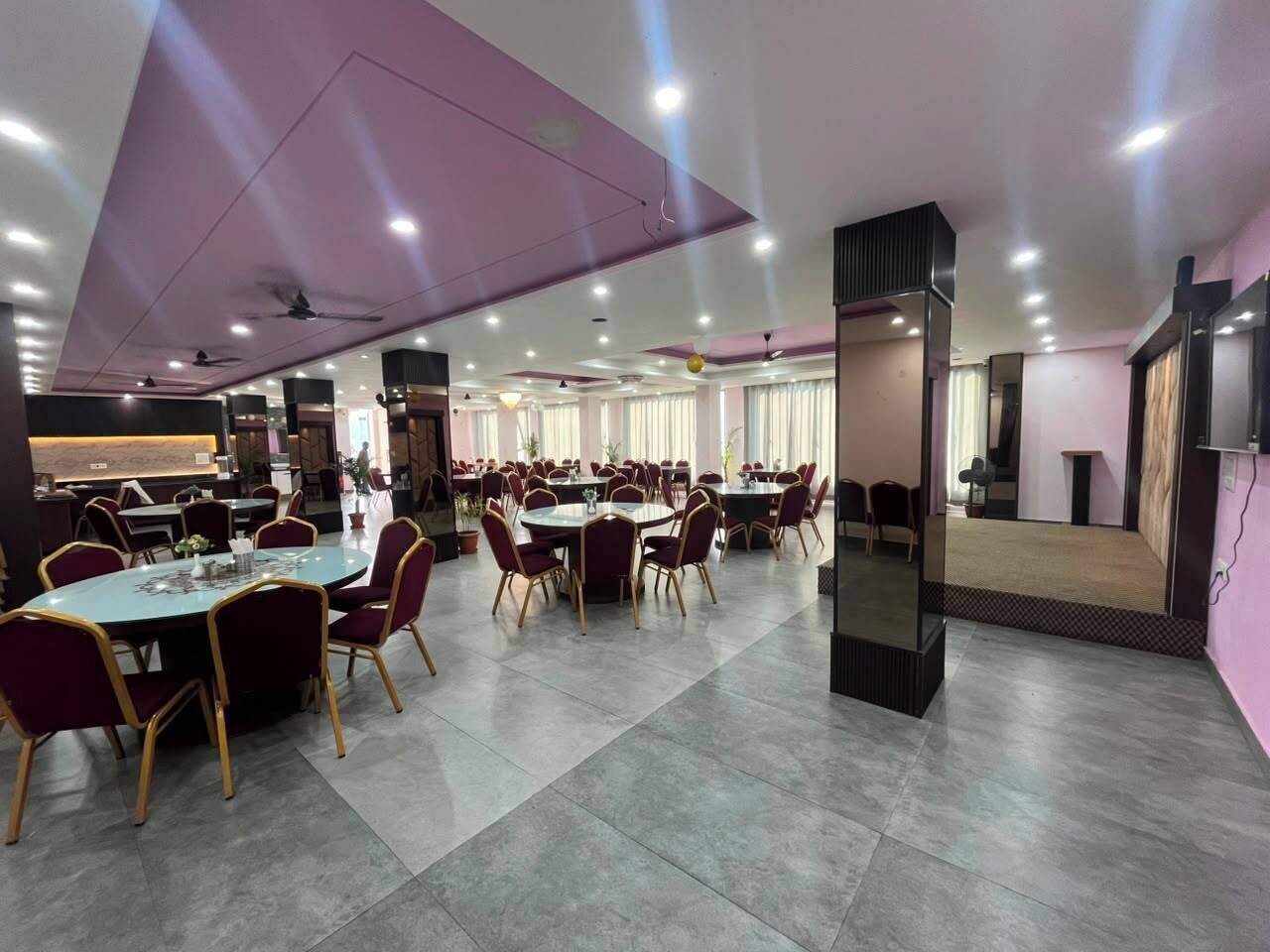 A spacious dining area with multiple round tables and chairs, a modern ceiling design, and a large mirror on one wall.