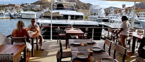 An outdoor dining area with tables and chairs, overlooking a marina with boats and a mountainous backdrop.