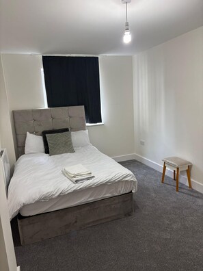 A bedroom with a bed, a window with dark curtains, a small wooden stool, and a ceiling light. - Central london 1-Bedroom Apartment with Garden View in London (London)