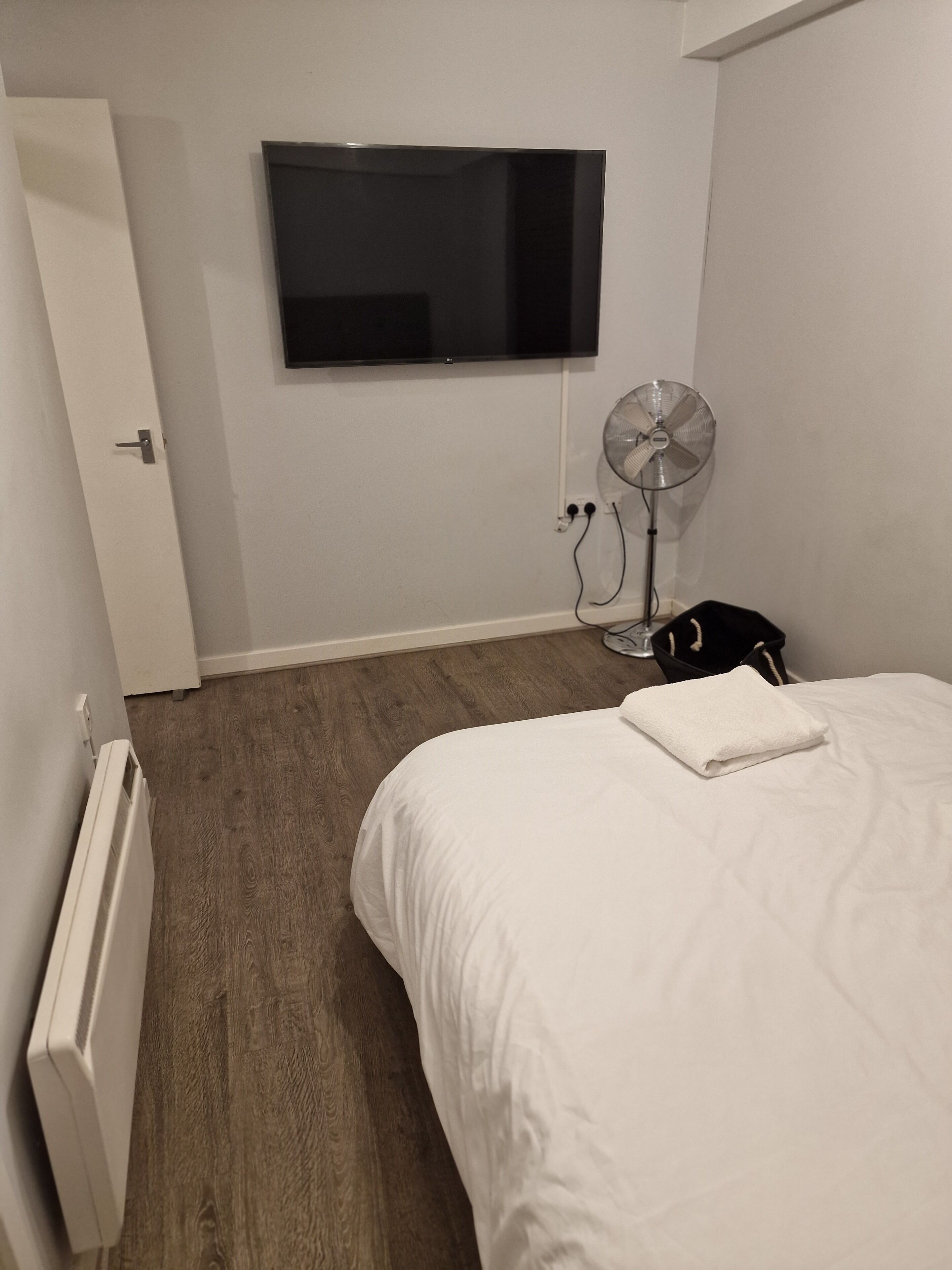 A bedroom with a bed, a TV mounted on the wall, a standing fan, and a radiator.