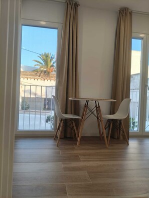 Una habitación con piso de madera, una mesa con sillas y ventanas grandes con cortinas.
