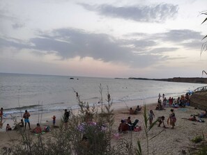Una playa con gente, algunos sentados y otros de pie, y un muelle al fondo.