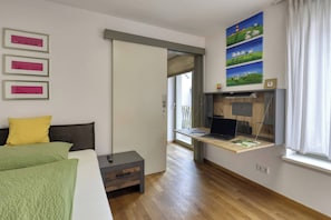 Ein Schlafzimmer mit einem Bett, einem Schreibtisch mit einem Laptop, einem Fenster mit Vorhängen und einer Tür zu einem anderen Raum.