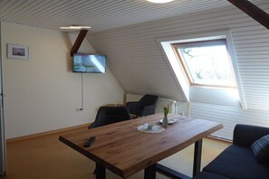 Ein Zimmer mit einem Holztisch, schwarzen Stühlen, einem an der Wand montierten Fernseher und einem Fenster mit Blick auf Bäume.