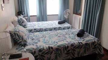 Una habitación de hotel con dos camas, una mesita de noche con lámpara, un ventilador y una ventana con cortinas.