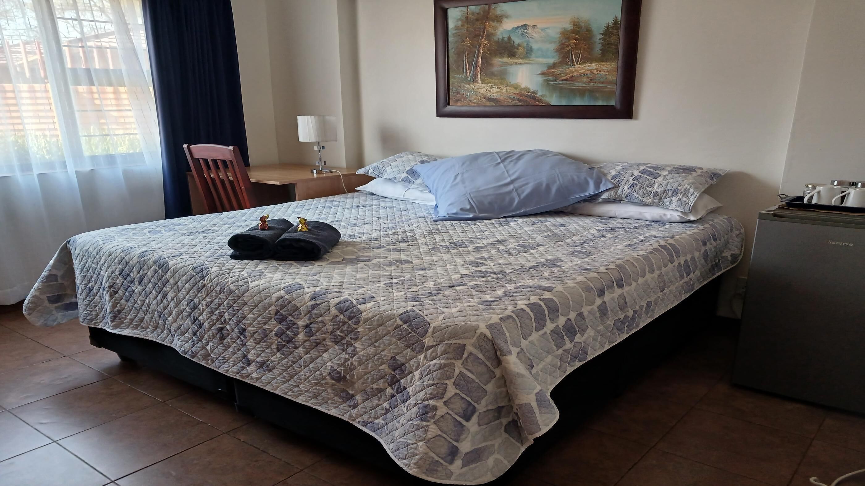 A hotel room with a bed, a painting on the wall, a desk with a lamp, and a refrigerator.