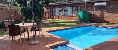 A swimming pool with a brick deck, a table with chairs, and a large umbrella.
