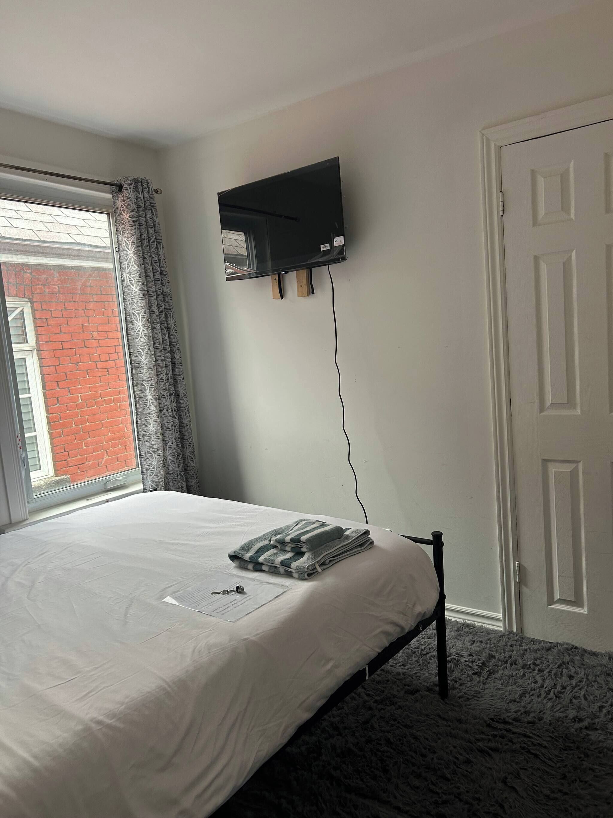 Une chambre à coucher comprenant un lit, un téléviseur à écran plat fixé au mur, une fenêtre avec des rideaux et une porte.