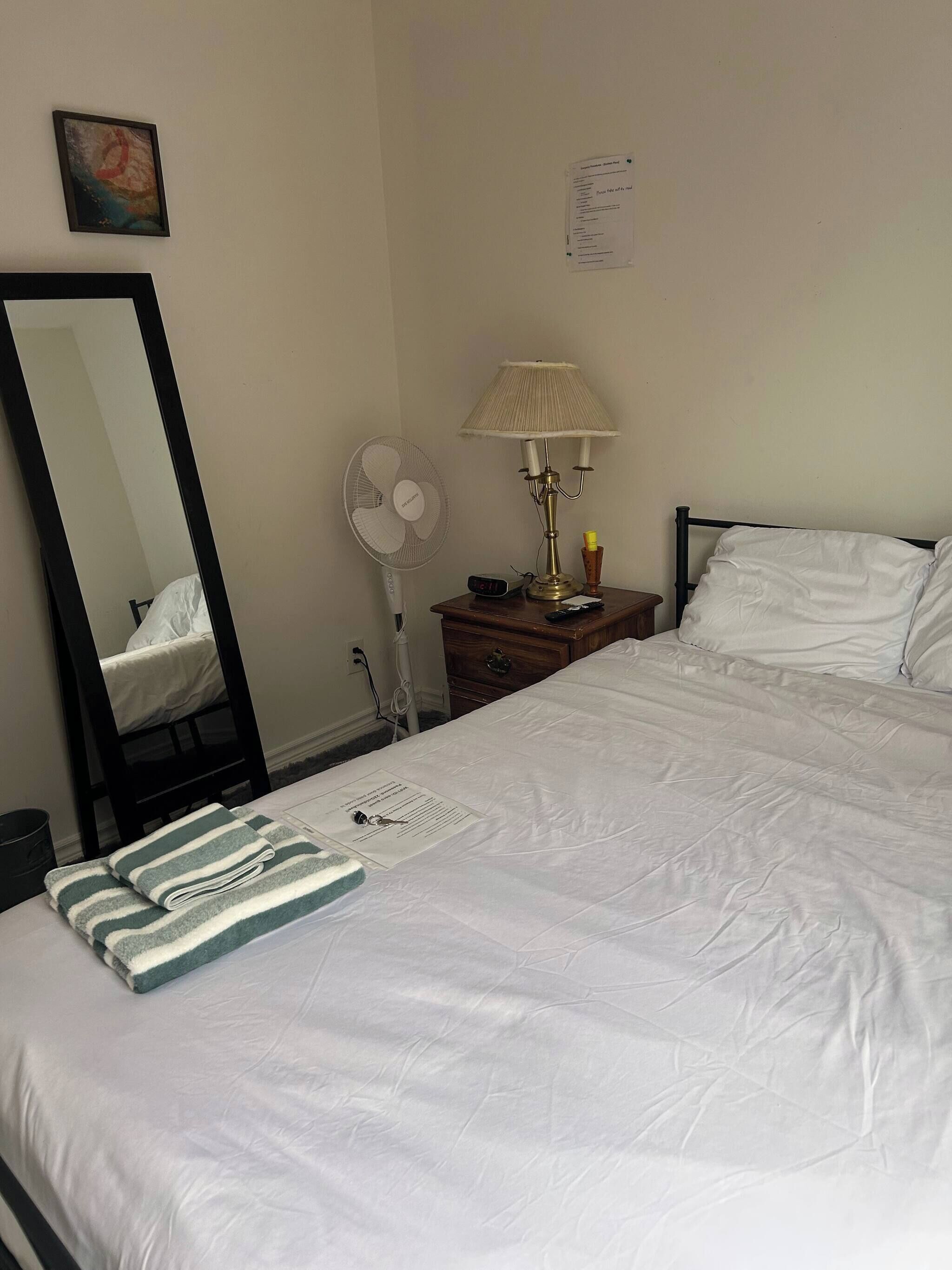 A neatly made bed with white bedding, a bedside lamp, a fan, and a mirror on the wall.