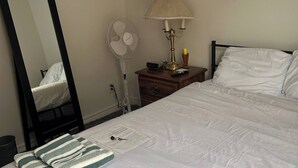 Una cama bien hecha con ropa blanca, una lámpara de noche, un ventilador y un espejo en la pared.
