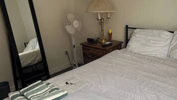 A neatly made bed with white bedding, a bedside lamp, a fan, and a mirror on the wall.