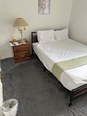 A bedroom with a bed, a nightstand with a lamp, a small wooden dresser, and a grey shaggy carpet.