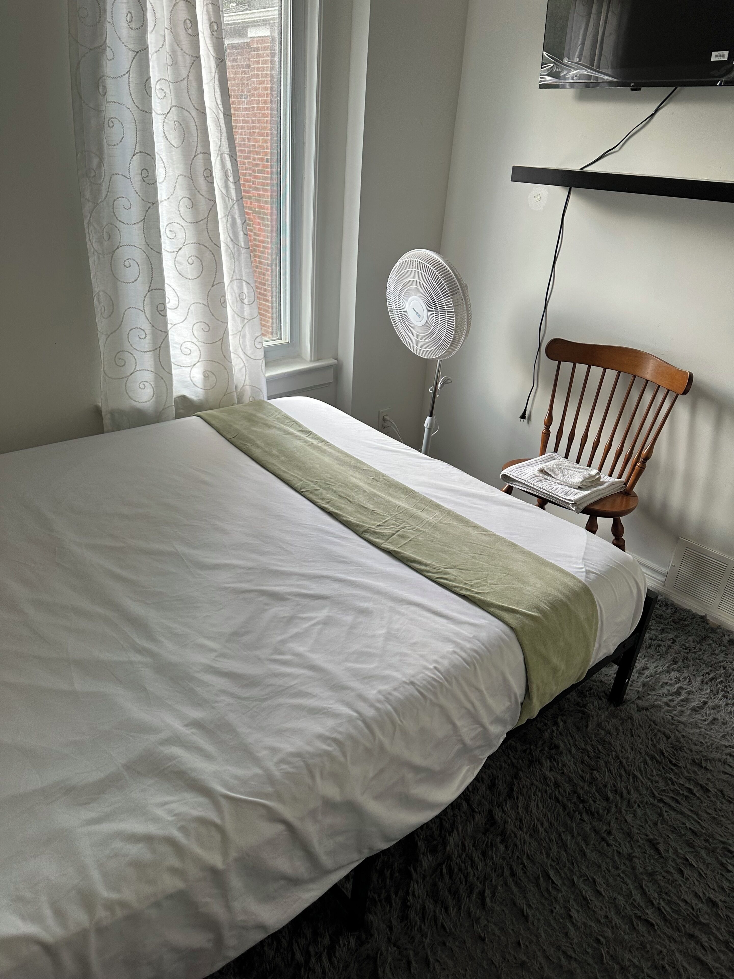 Un lit bien fait, avec un plaid vert, un ventilateur sur pied, une chaise en bois et un téléviseur fixé au mur.