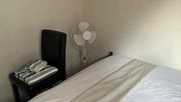 Una stanza essenziale con un letto, una sedia, una ventola a piantana e un oggetto fissato alla parete.