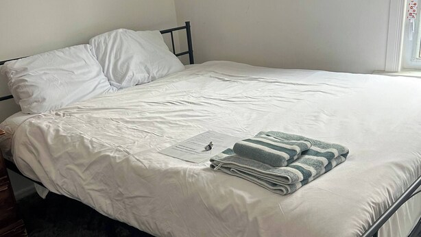 Una cama con ropa de cama blanca, estructura de metal oscura y una manta a rayas grises y blancas.