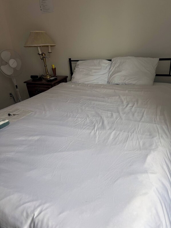 A bed with white linens, a bedside table with a lamp, and a wall-mounted fan.