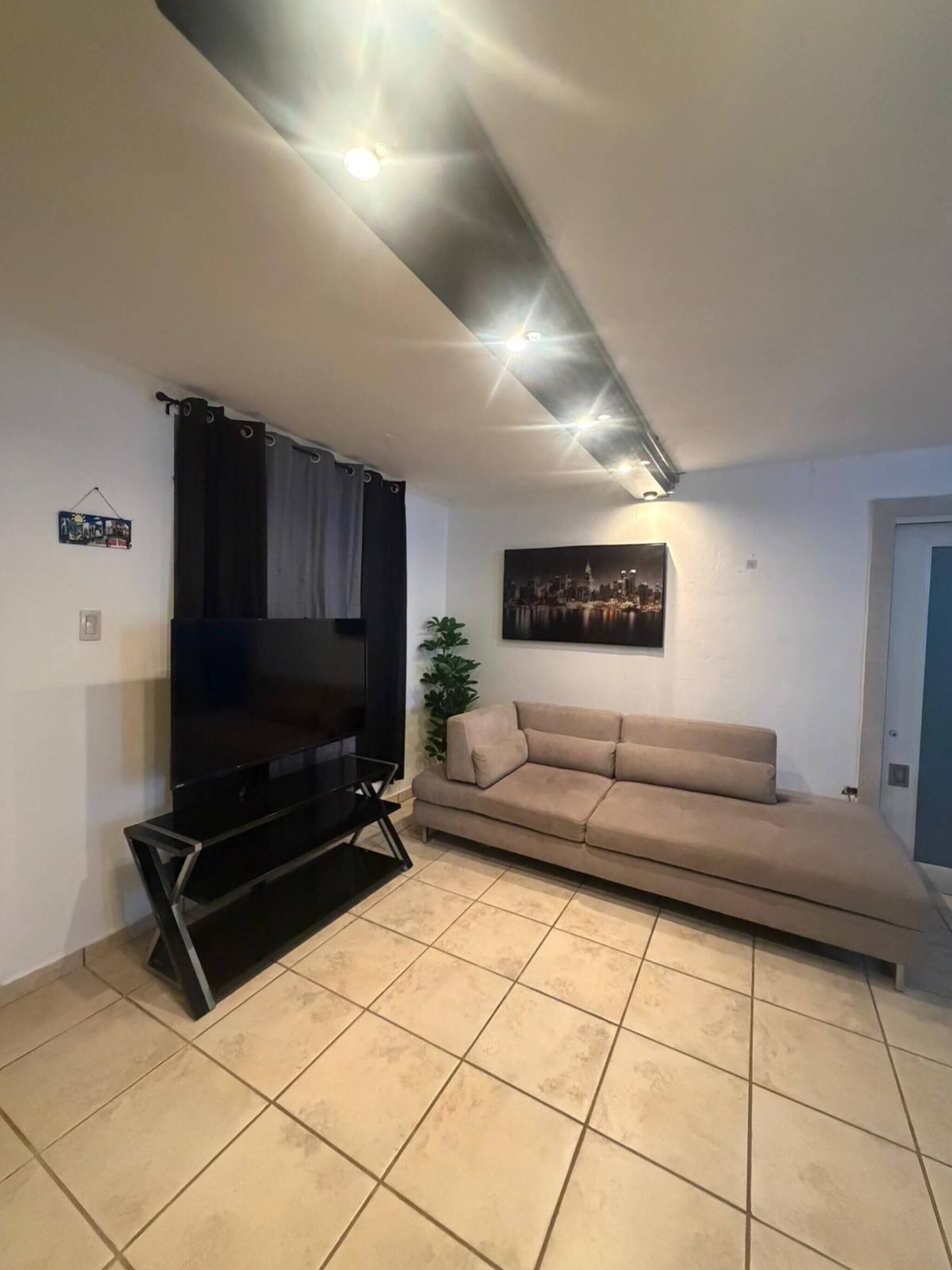 Una sala de estar con una televisión de pantalla plana, un sofá color beige, un cuadro de un paisaje urbano e iluminación empotrada en el techo.
