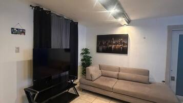 Una sala de estar con una televisión de pantalla plana, un sofá color beige, un cuadro de un paisaje urbano e iluminación empotrada en el techo.