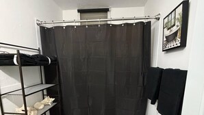 Bany compacte amb vàter, cortina de bany, paraigua de tovalloles i prestatgeries amb productes de tocador.