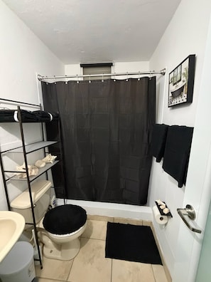 A compact bathroom with a toilet, shower curtain, towel rack, and a shelf with toiletries. - Your Adventure Home El Yunque, Culebra & Icacos (Luquillo city)