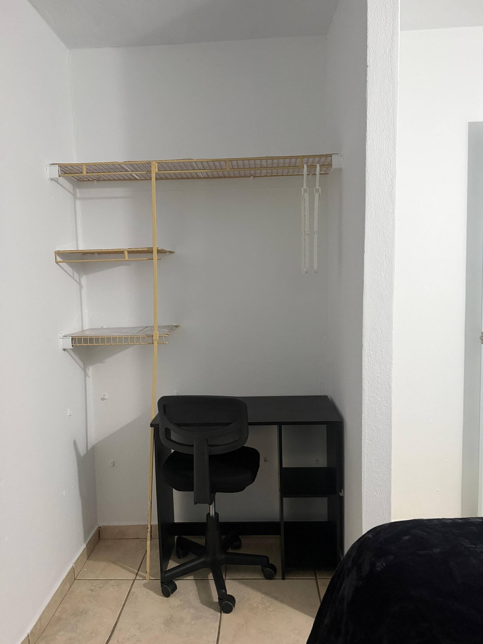 A corner room with a black office chair, a desk, and a white wall with a metal shelf unit.