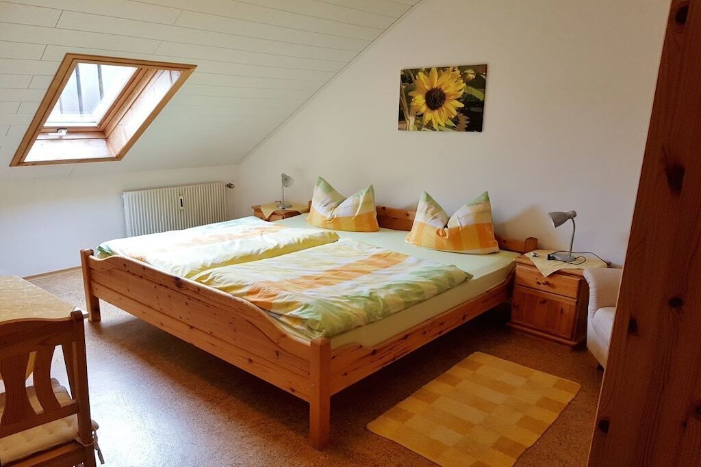 Ein Schlafzimmer mit schrägen Wänden, einem Bett mit gestreifter Bettwäsche, einem Holz-Kopfteil, einem Nachttisch, einem Sessel, einem Heizkörper und einem Gemälde einer Sonnenblume an der Wand.