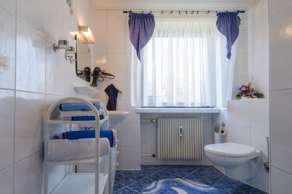 Ein Badezimmer mit einem weißen Waschbecken, einer Toilette, einem Handtuchhalter mit gefalteten Handtüchern, einem Fenster mit Vorhängen und einem Heizkörper.