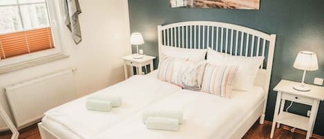 A neatly made bed with pillows and a blanket, a bedside table with a lamp, and a window with blinds.