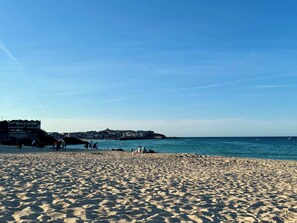 Ein sandiger Strand mit Fußspuren, die zum Wasser führen, ein klarer Himmel und Gebäude im Hintergrund.
