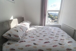 A bed with a patterned bedspread, a window with a view of the outside, and a radiator on the wall. - Seafront Soleil (Penzance)