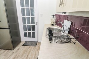 A kitchen with a stainless steel refrigerator, a door with frosted glass panels, a black dish rack, and a tiled backsplash. - Seafront Soleil (Penzance)
