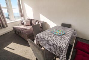 A room with a sofa, a table with a patterned tablecloth, and a window with curtains. - Seafront Soleil (Penzance)