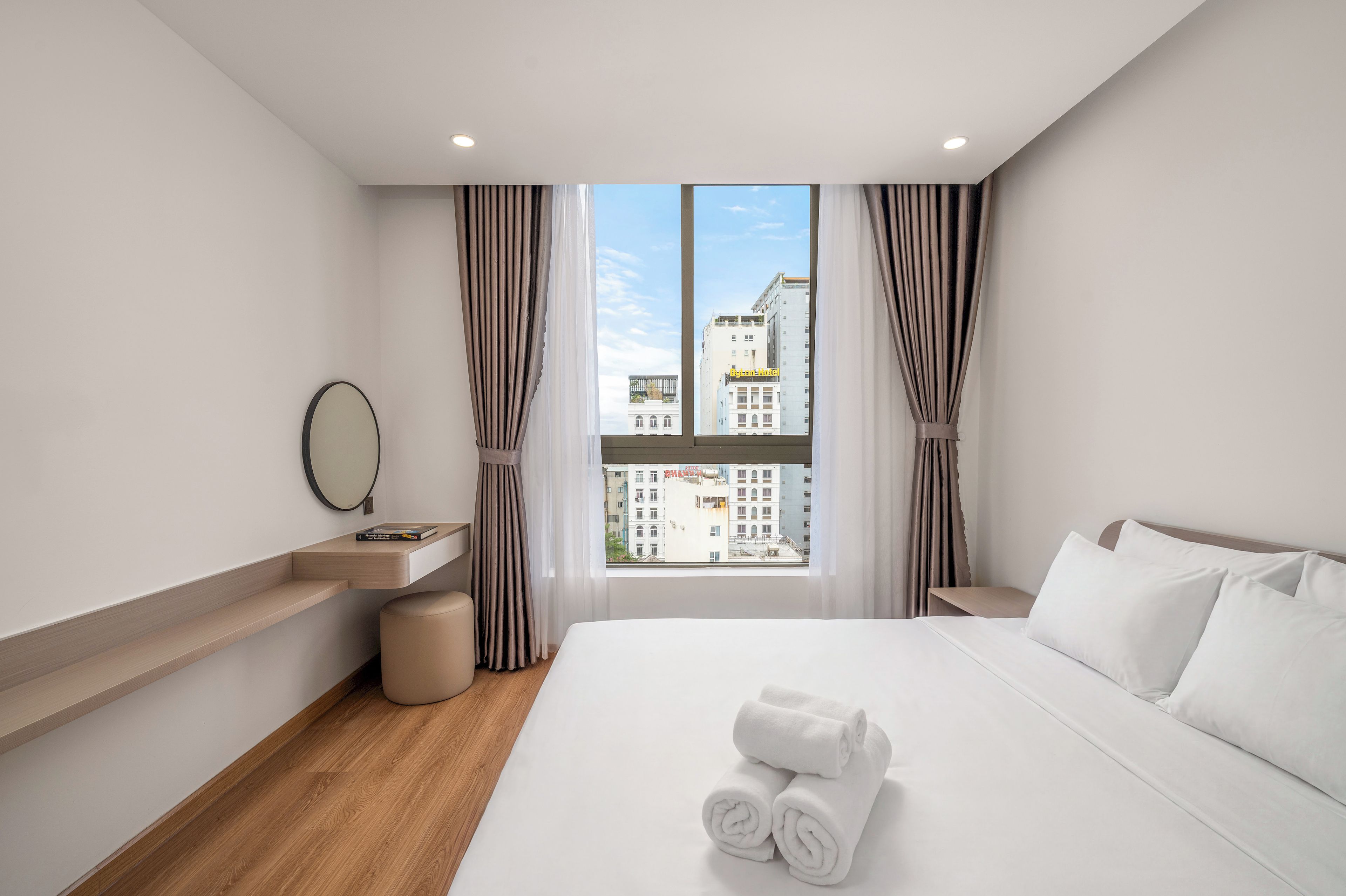 A hotel room with a bed, a bedside table, a mirror, and a view of buildings outside the window.