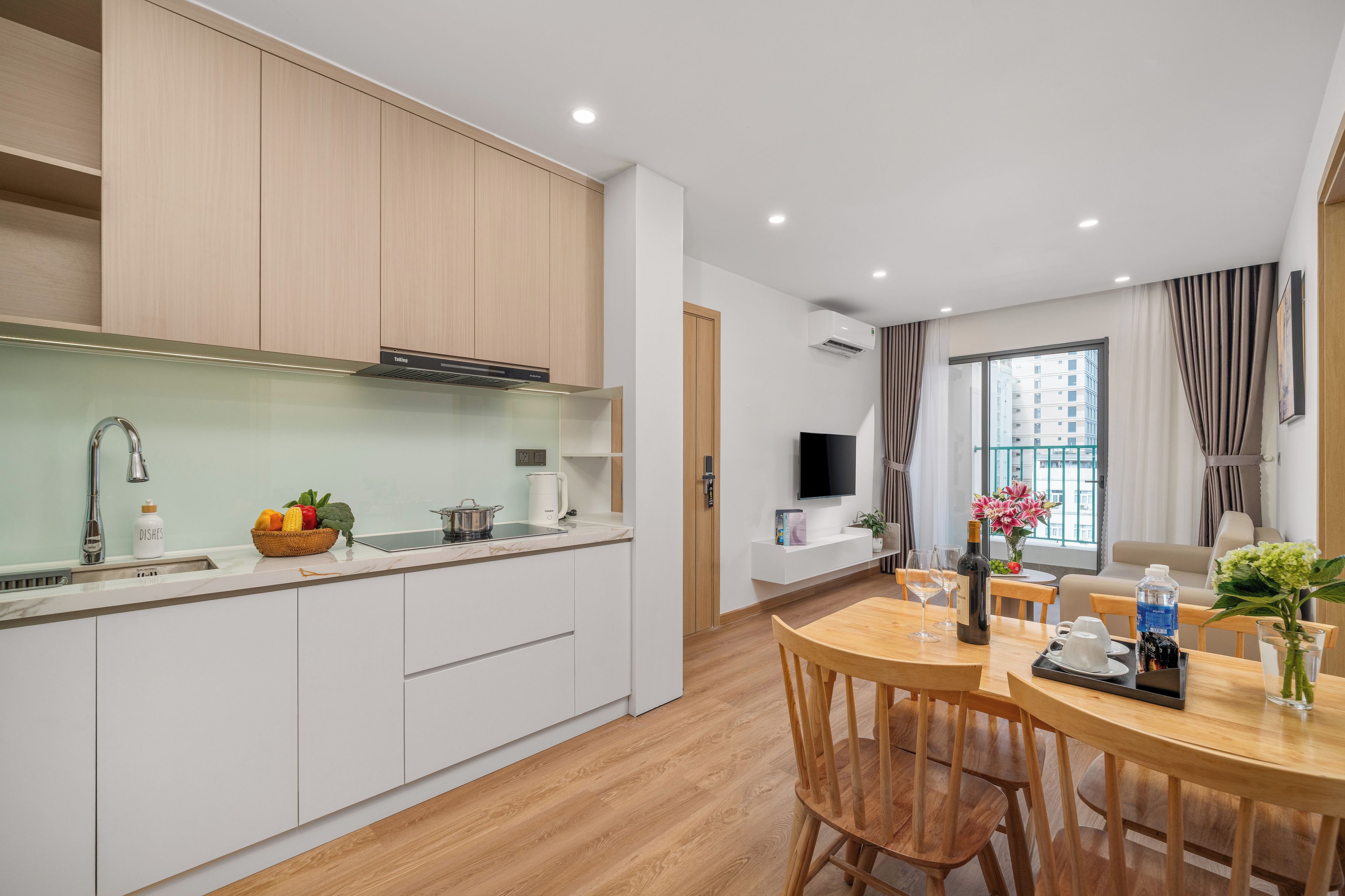Una cocina moderna con armarios blancos, una zona de comedor con mesa y sillas de madera, un televisor montado en la pared y una ventana con cortinas.