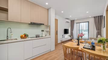 Una cocina moderna con armarios blancos, una zona de comedor con mesa y sillas de madera, un televisor montado en la pared y una ventana con cortinas.