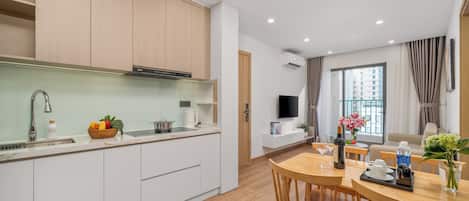 A modern kitchen with white cabinets, a dining area with a wooden table and chairs, a TV mounted on the wall, and a window with curtains.