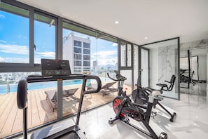 Un gimnasio moderno con equipo de ejercicio, una zona junto a la piscina y vistas a la ciudad.