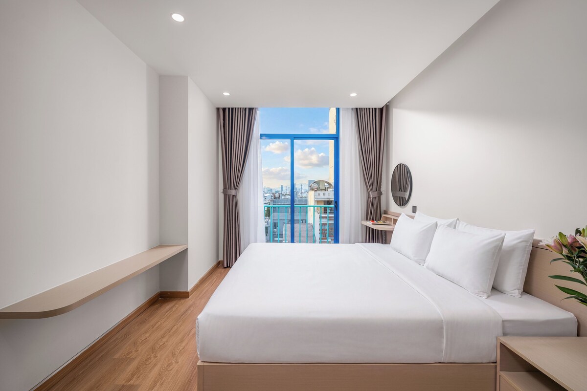 Habitación de hotel moderna con una cama grande, una mesita de noche, un escritorio pequeño y vistas de la ciudad a través de un ventanal amplio.
