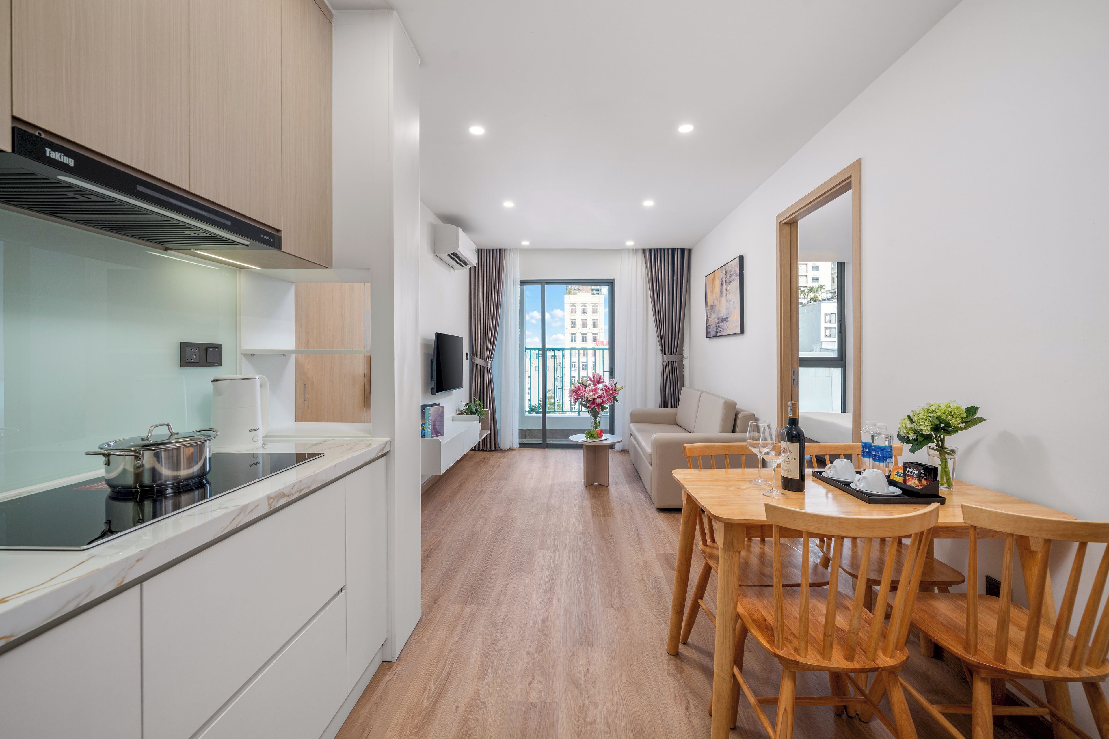 Uma cozinha moderna com fogão embutido, uma sala de jantar com mesa e cadeiras de madeira, e uma sala de estar com sofá e televisão.
