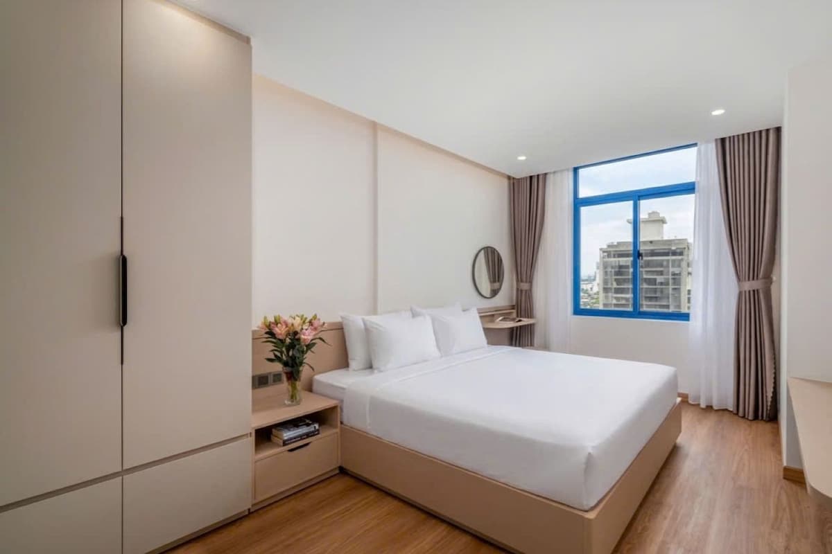 A modern bedroom with a large bed, a wardrobe, a bedside table with a vase of flowers, and a window with a view of buildings.