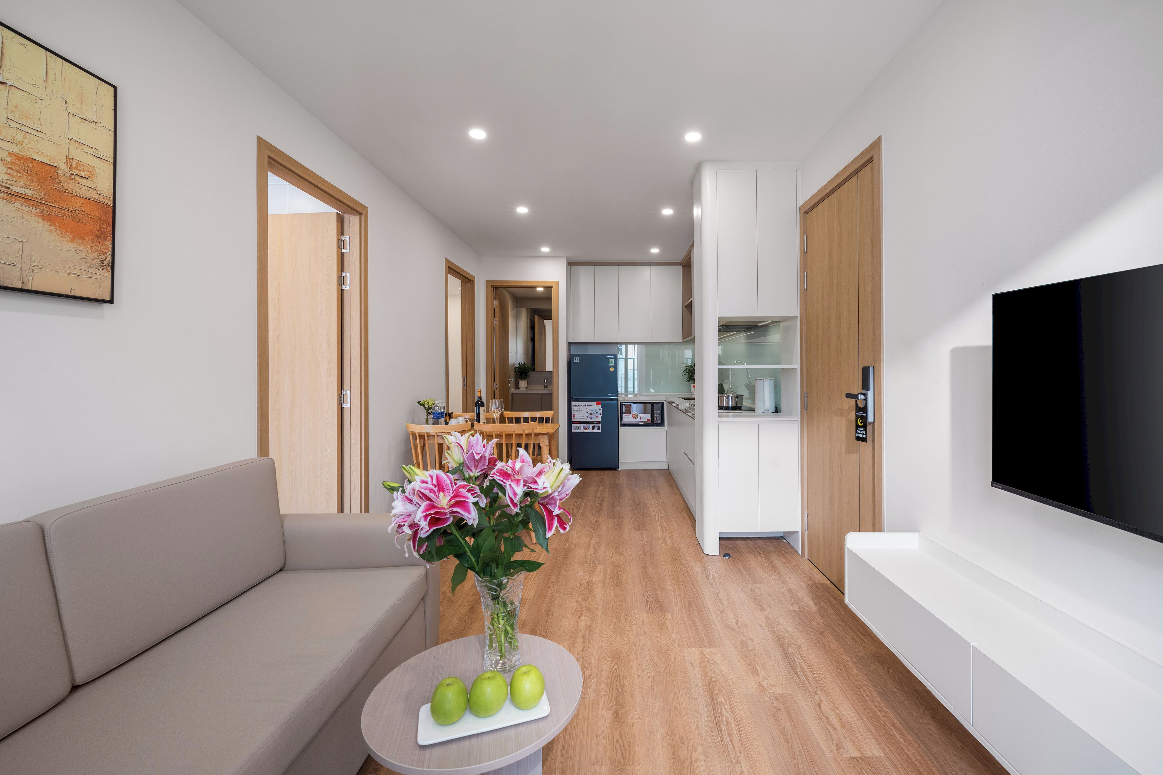 A modern living room with a sofa, a coffee table with apples and flowers, a TV, and a kitchen area in the background.