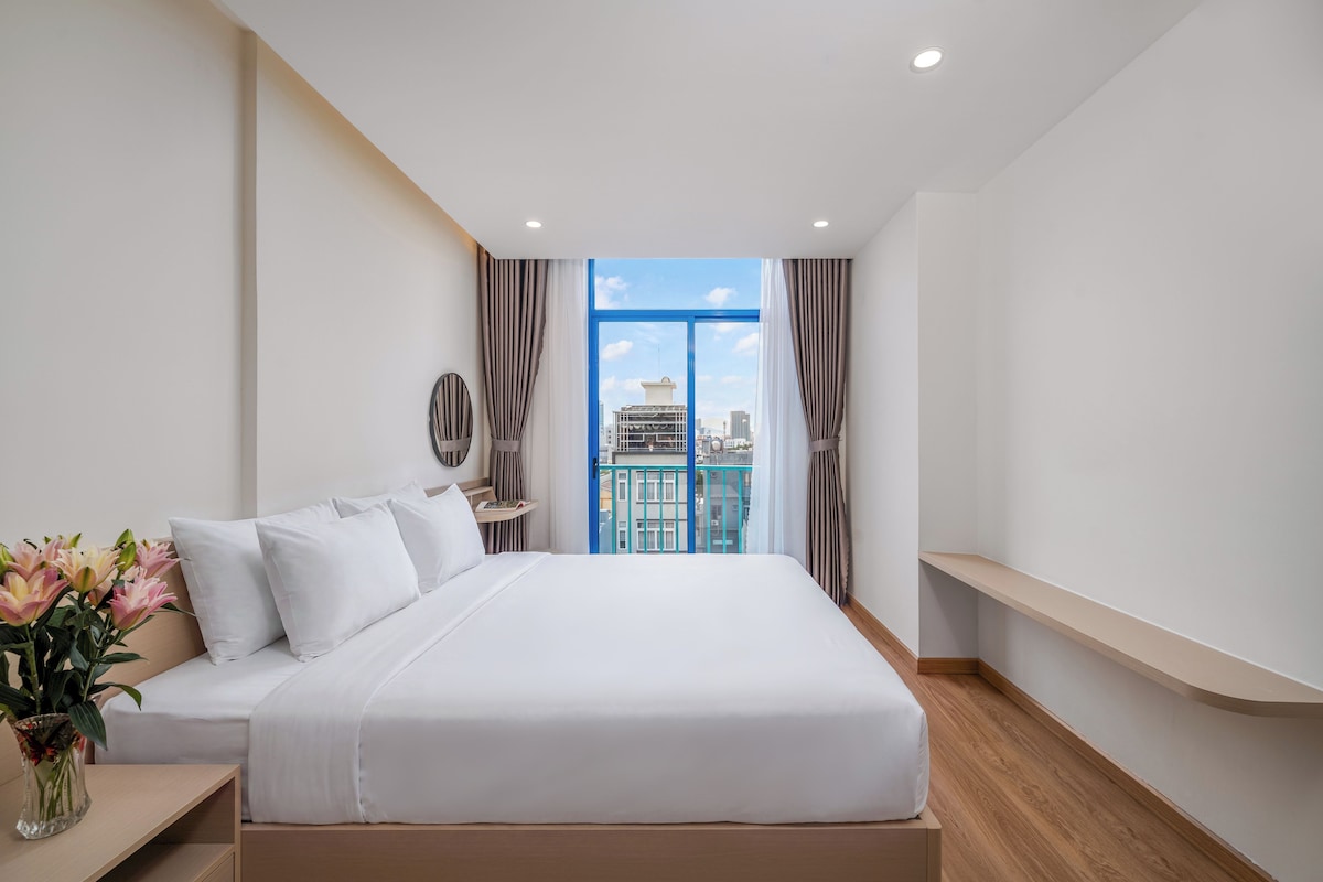 Habitación de hotel con una cama grande, una mesita de noche, un jarrón con flores y vistas a la ciudad a través de un ventanal amplio.