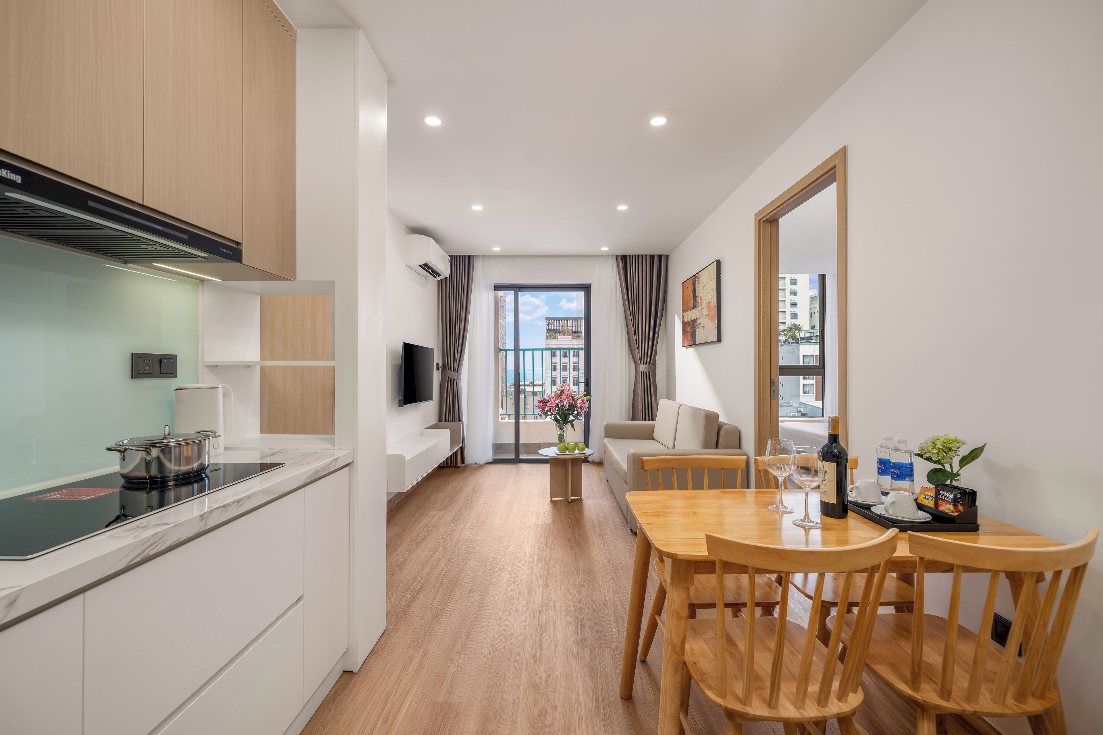 A modern kitchen with a built-in stove, a dining area with a wooden table and chairs, and a living area with a sofa and a TV.