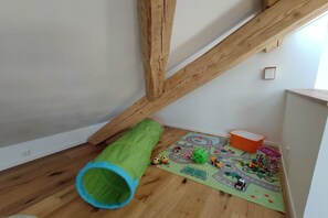 A room with a wooden beam ceiling, a green tunnel play structure, a colorful play mat, and a small orange container. - Holiday apartment in Görisried with Wi-Fi (Görisried)